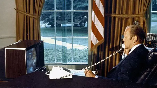 ABD Başkanı Gerald Ford, 1975 yılında Oval Ofis'teki masasında oturmuş, Apollo uzay gemisindeki astronotlarla telefonda konuşuyor. Konuşurken önündeki monitörü izliyor.
