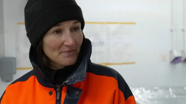 Liz Thomas, turuncu ve siyah koruyucu bir kıyafet ve şapka giymiş bir şekilde bir laboratuvarın içinde duruyor. Arka planda duvarda bir tabela ve el dezenfektanları var.