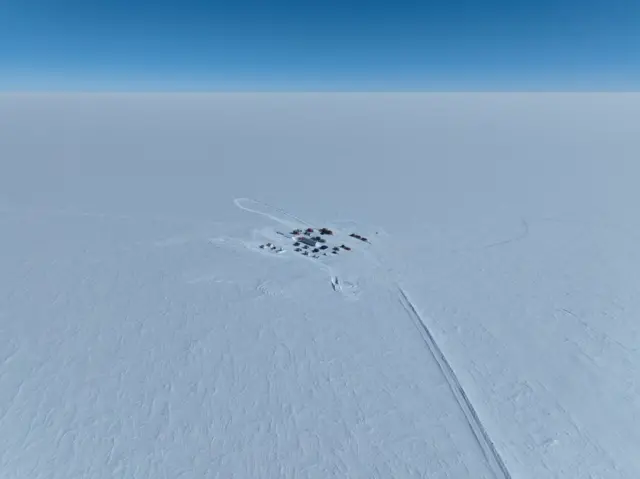Parlak beyaz Antarktika buz tabakasının ortasında küçük bir çadır ve geçici barınaklar görünüyor. Kamptan uzanan, ulaşım yolunu andıran, dağılmış kar izleri. Gökyüzü parlak mavi.