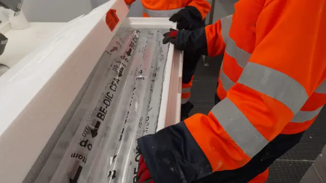 Turuncu ve siyah koruyucu kıyafet ve eldiven giymiş bir kişi, ellerini plastik ambalajla kaplı uzun buz tüplerinin üzerine koymuş. Tüpler büyük bir kutunun içinde.