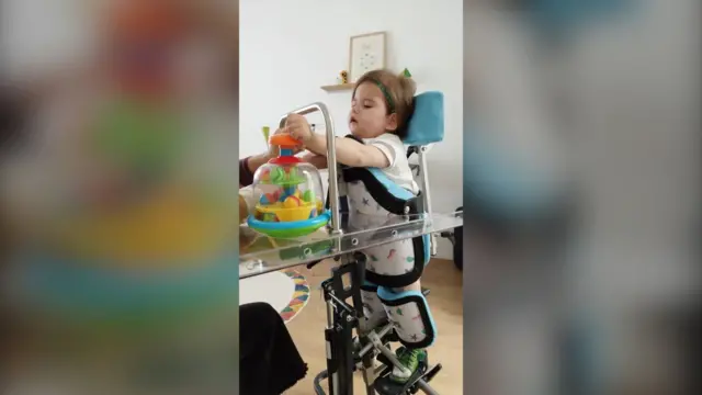 Laia, dik durup oyuncaklarıyla oynamasını sağlayan metal bir yapıya bağlı haldeyken. 